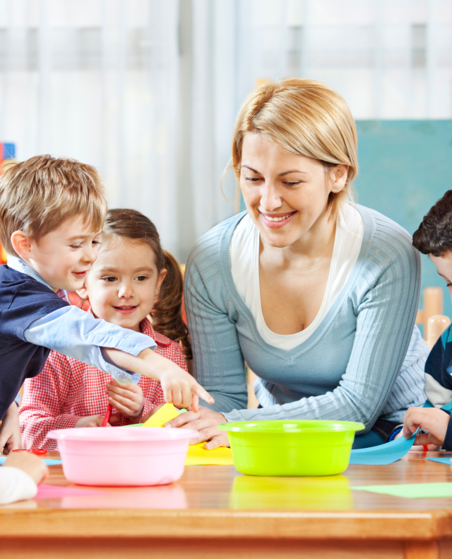Afbeelding van juf die met 4 kinderen aan een tafel zit met 2 bakjes en gekleurd papier